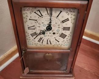 A wooden-framed wall clock featuring a cream-colored face with Roman numerals and ornate hands. The case appears to be made of stained wood with a glass panel below the clock face.
