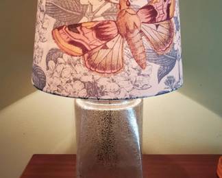 Table lamp with a cylindrical frosted glass base and a fabric shade printed with colorful seashell and marine motifs. The shade features pink, orange, and blue tones with a vintage-inspired design.