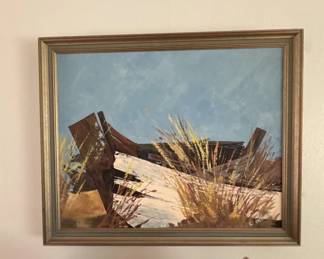 Framed print or painting depicting a rustic wooden structure or debris in a desert or arid landscape with golden grasses and blue sky. The piece is mounted in a wooden frame with a neutral finish.