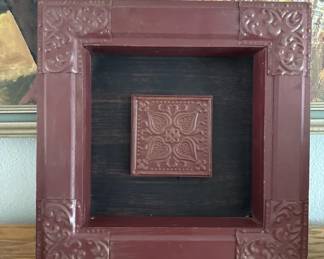 Square wooden frame with burgundy finish and ornamental carved corners, featuring a small embossed copper or bronze tile in the center against a dark background. A simple shadow box-style display piece.