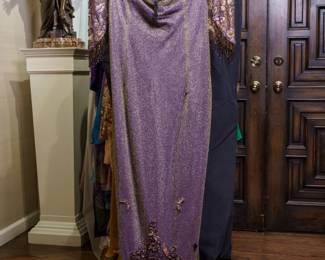 LOT 52 – Purple Silk Satin and Iridescent Gold Lace Evening Gown, with Silk Satin Floral Sleeves, and Beaded Embellishments. A stunning lavender evening gown crafted from luxurious purple silk satin, layered with iridescent gold lace for a beautiful color-shift effect that gives the gown a shimmering lavender appearance. Elegant floral silk-satin sleeves fall gracefully to the elbow and feature exquisite hand-sewn beadwork, from the neckline and swirling floral designs on the sleeves to delicate beaded details cascading from the sleeve edges. Designed by famous fashion and costume designer Margi Kent. Sold as a full ensemble set. • Approx. Size ~8 to 8/10; Approx. Measurements: Waist: 30”, Bust: 36, Hips: 36. • Owner’s note: the gowns can be adjusted in size; there is plenty of fabric in the seams.
