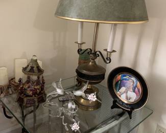 Vintage brass table lamp with a green shade, displayed on a glass-top end table alongside a decorative collector plate, a glass bird figurine, and a small carousel music box.