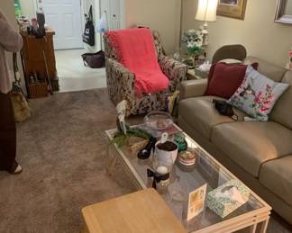 Living room furniture set including a beige leather sofa, a glass-top coffee table with a white frame, and a patterned armchair draped with a pink throw blanket.