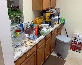 Assorted kitchenware and household items, including glassware, ceramic canisters, a Betty Crocker slow cooker in its box, and a grey step-on trash can, arranged on and around oak kitchen cabinets.