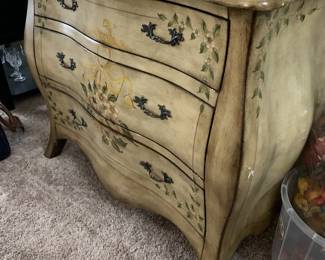 French Provincial style bombee chest of drawers featuring a hand-painted floral motif, antiqued cream finish, and ornate metal pulls.