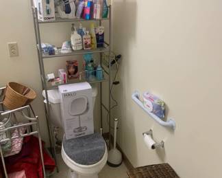 Metal over-the-toilet shelving unit holding various toiletries, a Conair fabric steamer, and an iron, alongside a boxed Conair Halo lighted mirror resting on the toilet seat.