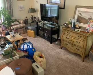 Living room filled with various items for sale, including a flat-screen TV on a black stand, a hand-painted French provincial style chest of drawers, framed wall art, and assorted home decor.