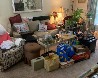Living room furniture and decor, including a beige leather sofa, floral armchair, brown ottoman, glass-top coffee table, and various household items.