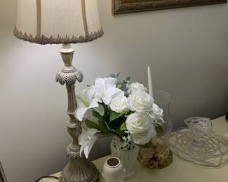 Traditional table lamp with an ornate, distressed-finish base and a fringed fabric shade, shown alongside a faux floral arrangement in a white vase and other tabletop decor.