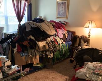 Large assortment of women's clothing piled on a bed, alongside bedroom furniture, a brass table lamp, and framed wall art.