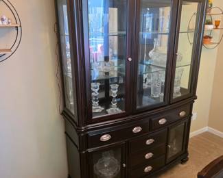 dark wood china hutch with glass display doors, interior lighting, and lower storage drawers, filled with cut crystal items.