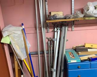 Collection of long metal bar clamps leaning against a pink wall alongside cleaning tools, shelving, and a blue painted tool chest.