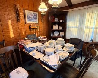 A dining table and dark wood corner hutch filled with floral dinnerware, stackable dish sets, linens, and home decor items in a wood-paneled room.