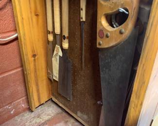 Collection of vintage hand saws hanging on a pegboard inside a wooden cabinet, including Japanese pull saws and a traditional hand saw.