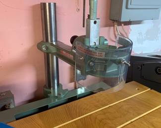 Shopsmith router arm attachment mounted on a wooden table, featuring a clear safety guard and sage green cast iron body.