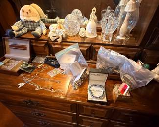 Collection of assorted costume jewelry, glass figurines, cut crystal glassware, and small collectibles displayed atop a dark wood dresser.