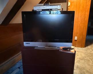Black and silver flat-screen television with a remote control, sitting on a dark wooden Media cabinet alongside a DVD player.