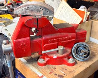 Red Craftsman bench vise with swivel base, mounted on a wooden workbench in a workshop setting.