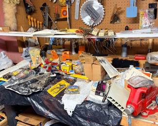 Workshop workbench cluttered with assorted hand tools, hardware, supplies, and a red bench vise, framed by pegboard storage.