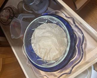 Collection of kitchenware stored in a cabinet drawer, including stacked glass serving bowls, plastic measuring cups, and a food processor attachment.