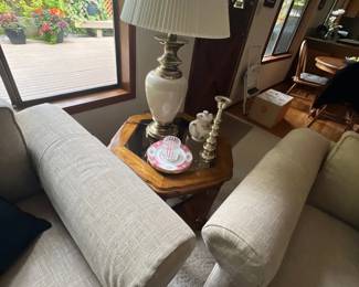 A vintage table lamp with a pleated shade and brass-tone accents sits on a hexagonal wood end table between two beige upholstered armchairs. Decorative items on the table include a small teapot, candlestick holder, and trinket dish.