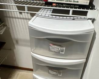 Sterilite white plastic three-drawer storage cart, positioned below a wire shelf holding a collection of paperback and hardcover books.