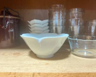 Collection of kitchenware, including a white porcelain flower-shaped bowl, clear glass dessert dishes, and a brown ceramic crock on a wooden shelf.