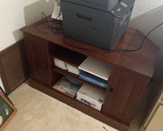 Brother all-in-one laser printer sitting atop a dark wood corner media cabinet or office console with open shelving and side doors.
