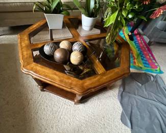 Hexagonal wood and glass coffee table displaying decorative orb centerpiece, potted houseplants, and a floral arrangement.