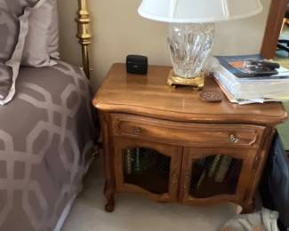 French Provincial style wooden nightstand featuring a top drawer, double cabinet doors with wire mesh inserts, and a crystal table lamp with a pleated shade.