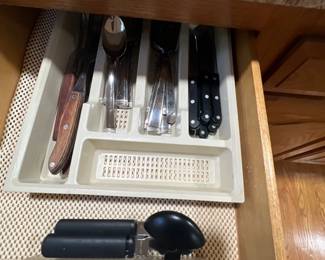 Kitchen drawer containing an assortment of flatware, steak knives, a can opener, and bottle opener organized in plastic trays.