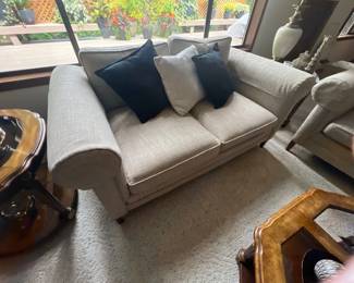 Contemporary beige upholstered loveseat featuring rolled arms and padded cushions, styled with black and patterned accent pillows.