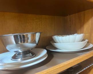 Selection of serveware including a silver-tone footed bowl on white porcelain plates and stacked fluted white ceramic dishes.