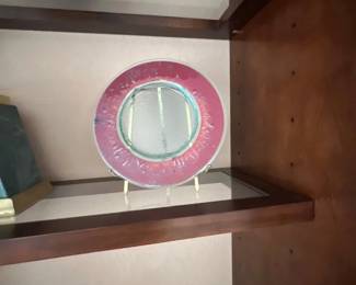 Round decorative glass plate with a textured magenta rim and clear center, displayed on a small brass stand within a wooden shelf.