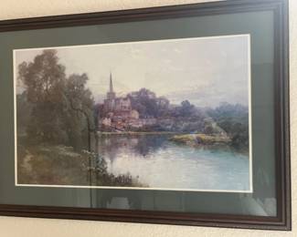 Framed landscape art print featuring a European riverside village with a church spire, housed in a dark wooden frame with a green mat board.