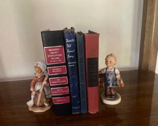 Collection of vintage hardcover books flanked by two ceramic Hummels child figurines on a wooden shelf.
