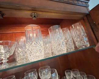 Collection of crystal highball glasses and stemware featuring crosshatch cut details, arranged inside a lighted wooden china cabinet.