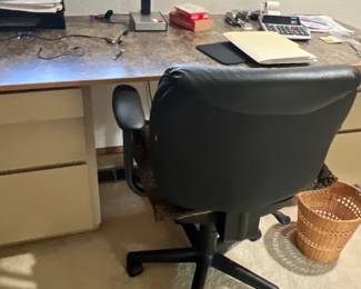 Home office setup featuring a large laminate-top desk, a black faux leather rolling desk chair, a vintage fluorescent desk lamp, and various office accessories.