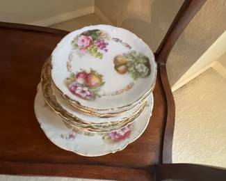 Stack of vintage hand-painted porcelain plates with floral and fruit motifs and gold rim accents, resting on a wooden shelf.