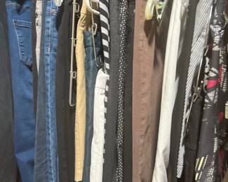 Closet rod filled with an assortment of women's pants, including blue denim jeans, khaki trousers, and patterned slacks on plastic hangers.