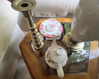 Collection of tabletop decor including a brass candlestick, porcelain teapot, decorative saucers, and a lamp resting on a hexagonal glass-top side table.