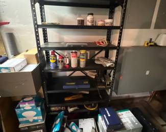 Black metal industrial shelving unit holding various workshop chemicals, lubricants, and hardware, surrounded by boxed lighting supplies.