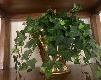 Faux green ivy plant arranged inside a polished brass bucket-style planter with a pedestal base, displayed on a wooden shelf.