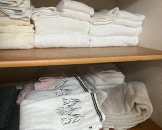 Assorted stacked bath towels and hand towels, including white terrycloth pieces and embroidered designs, arranged on wooden linen closet shelves.