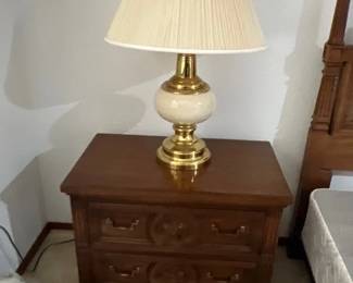 Traditional dark wood two-drawer nightstand with carved detailing, topped with a polished brass and ceramic table lamp with a pleated shade.