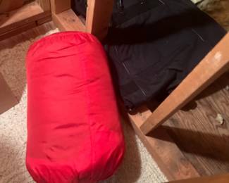 Bright red rolled sleeping bag packed in its matching stuff sack, stored alongside a black garment bag under wooden framing.