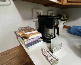 Black Mr. Coffee 12-cup drip coffee maker sitting on a kitchen counter alongside a stack of cookbooks and kitchen towels.