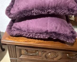 Pair of purple damask throw pillows with brush fringe trim stacked on a vintage wood nightstand.