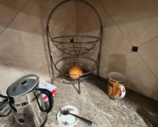 Two-tier metal wire fruit basket shown alongside a stainless steel electric milk frother and kitchen accessories on a granite countertop.