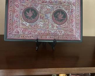 Hand-carved Olinalá lacquered wooden tray from Guerrero, Mexico, displayed on a stand. Features intricate etchings of birds, flora, and fauna in shades of red and white on a dark background.